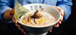 Kanada-Ya | Ramen Near Me | A person in a blue shirt holds a bowl of ramen with sliced meat, seaweed, spring onions, and broth. The white bowl has black and gold characters on it. | London
