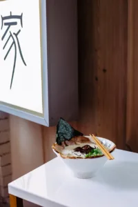 Kanada-Ya | Ramen Near Me | A bowl of ramen with sliced pork, seaweed, spring onions, and chopsticks rests on a white table beside a wooden wall and a large sign with black Japanese characters. | London