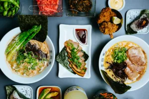 Kanada-Ya | Ramen Near Me | A top-down view of a table set with two bowls of ramen, side dishes including fried chicken, edamame, pickled vegetables, rice balls, dipping sauce, and a drink. The ramen bowls are garnished with spring onions and seaweed. | London