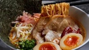 Kanada-Ya | Ramen Near Me | A close-up of a bowl of ramen with sliced pork, soft-boiled eggs, noodles held by chopsticks, bean sprouts, spring onions, pickled ginger, seaweed, and a dollop of red chilli paste in rich broth. | London